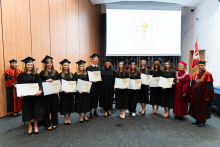 A group of about a dozen people. From the left stand pharmacy graduates dressed in togas. In their hands they hold folders with diplomas spread out. On the right stands the vice-chancellor and the dean. They are all smiling. In the background, the WUM flag can be seen on the right, and on the left the pro-dean in a toga. A projection screen can also be seen behind the group.