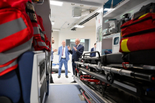 In the background, in the middle of the photo, there are three people in suits talking. The faces of these people are blurred. A stretcher with a black mattress is visible on the right. On the stretcher lies a red emergency bag. The interior of the ambulance is well lit by a white ceiling light.