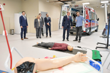 Five people standing in a room. An open ambulance can be seen in the background. The interior of the ambulance is clearly visible. In the foreground lies a mannequin on a black mat. The mannequin is dressed in a maroon sweatshirt and dark pants. Next to the mannequin is medical equipment, including a defibrillator. To the left, on a white table, lies an artificial hand with a bandage and traces of red dye.