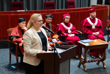 In the foreground, a middle-aged woman stands behind the lectern and speaks. She has glasses, light hair and wears a light jacket and dark dress. Behind her, the dean's and rector's authorities are seated on chairs.