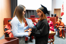 On the left is a woman in a blue blouse with long hair and glasses; on the right is a graduate in a black toga with red hair. The woman on the left hands the graduate her diploma. They shake hands in a gesture of congratulations. Other people can be seen in the background, the rector and dean authorities.