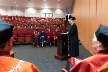 A young man dressed in an academic toga - a representative of the graduates - stands behind the lectern and speaks. He is facing the photo, looking at the rector and dean authorities. Behind him, on red armchairs sit the participants of the ceremony.