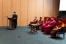 A young man, a representative of the local government, in a black toga stands behind a brown lectern and speaks. Behind him, the dean's and rector's authorities sit on chairs.