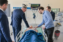 Four men standing around a hospital bed. On the bed lies a medical mannequin covered with a blue blanket. The men are dressed in suits and shirts. One of them points to the mannequin. The other is touching the mannequin's chest. A medical monitor with vital signs can be seen in the background. Next to it are medical devices.