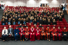 A large group of people in togas and birettas sit in the auditorium. In the first row, people in togas of different colors, behind them graduates with diplomas. The auditorium is full, there are people standing on the stairs on the sides.