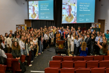 A group of people pose for a photo in an auditorium with red seats. In the background, two screens with the caption: 'CRAIG MELLO RNAi: Ancient Mechanism, New Medicines'. 
