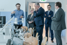 Five people dressed formally stand in a bright room with medical simulators. The walls are painted white and blue. In the foreground lie medical mannequins for dental training. The mannequins are on tables, and lamps hang above them. A door can be seen in the background.