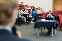 In the foreground, on the left, there is a person holding a microphone, whose face is blurred and invisible. In the center of the photo, a man is sitting at a table covered with a navy blue tablecloth, on which there are documents and a bottle of water. In the background, there is a group of people sitting on chairs, some of them holding notes or electronic devices.