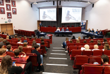 A large lecture hall with red seats, where a mock trial is taking place. At the front of the hall, there are two large screens displaying an image of a judge's gavel and the text “Mock Trial.” Several people are seated at tables at the front of the hall, representing various roles in the simulation. The audience consists of students.