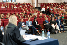 A lecture hall with red armchairs, where a mock trial is taking place. In the foreground, there is a table with a woman in a robe sitting at it—her face is not visible. In the background, there are a large number of participants sitting in armchairs, looking toward the podium.