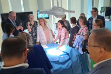  Grupa osób stoi wokół stołu operacyjnego w sali Centrum Symulacji Medycznych. Na stole leży niebieskie przykrycie chirurgiczne z narzędziami. Nad stołem lampa operacyjna, w tle dwa monitory i okna z żaluzjami. Twarze osób są zamazane, stroje różnorodne – formalne i codzienne.