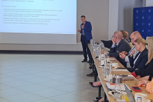Prezentacja w sali konferencyjnej. Mężczyzna w niebieskiej marynarce z mikrofonem wskazuje na ekran z tekstem o zmianach projektowych. Po prawej stronie osoby siedzą przy stole z wodą, filiżankami i notatkami, słuchając prezentacji.