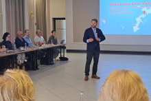 Mężczyzna w garniturze prowadzi prezentację przed grupą osób przy stole konferencyjnym. Na ekranie slajd z napisem „Średnia podaż kalorii 1768” i mapa świata z danymi o spożyciu kalorii. Sala jasna, z dużymi oknami i zasłonami po lewej stronie.