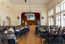 Duża sala wykładowa wypełniona uczestnikami siedzącymi na krzesłach i zwróconymi w stronę sceny. Na scenie znajdują się trzy osoby. 