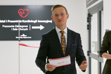 Mężczyzna w garniturze, prof. Marcin Grabowkski, trzyma kartkę papieru. Obok niego częściowo widoczna druga osoba. W tle czarny szyld z białym napisem: „Oddział i Pracownie Kardiologii Inwazyjnej, Pracownie Zabiegowe i Sale Chorych”. Na szyldzie czerwone serce z linią EKG i strzałka wskazująca kierunek.