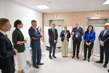 Osiem osób stoi w jasnym pomieszczeniu z białymi ścianami i sufitem. W tle dwie windy i białe parawany medyczne. Osoby ubrane elegancko, głównie w garnitury i marynarki. Po lewej stronie stół z butelkami wody mineralnej i kieliszkami.