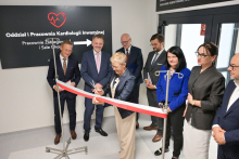 Grupa elegancko ubranych osób stoi w jasnym pomieszczeniu. Kobieta w środku przecina biało-czerwoną wstęgę nożyczkami. W tle czarna tablica z napisem „Oddział i Pracownia Kardiologii Inwazyjnej”, czerwoną grafiką serca z EKG i strzałką kierunkową.