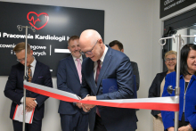 Mężczyzna w garniturze przecina czerwoną wstęgę. Obok stoją formalnie ubrane osoby. W tle czarna tablica z białym napisem „Pracownia Kardiologii” i czerwonym sercem z linią EKG. Po prawej stronie ściany widoczny napis „ODDZIAŁ PRACOWNIE HEMODYNAMIKI”. Pomieszczenie jest jasne.