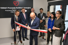 Mężczyzna w garniturze przecina biało-czerwoną wstęgę podczas otwarcia oddziału. W tle czarna tablica z białym napisem: „Oddział i Pracownia Kardiologii Inwazyjnej, Pracownie Zabiegowe i Sale Hybrydowe”. Obok elegancko ubrani uczestnicy, część trzyma dokumenty.
