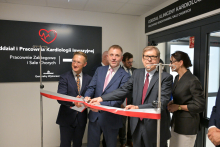 Trzech mężczyzn w garniturach przecina czerwoną wstęgę. W tle czarna tablica z białym napisem „Oddział i Pracownia Kardiologii Inwazyjnej, Pracownie Zabiegowe i Sale Chorych” oraz czerwonym sercem z linią EKG. Po prawej wejście z napisem „Oddział Kliniczny Kardiologii, Pracownie Hemodynamiki, Sale Chorych”.