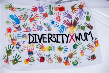 White banner covered with colorful handprints in the colors of red, blue, green, yellow and purple. In the center you can see the inscription “DIVERSITY X WUM”, where the letter “X” is red and the others are black.