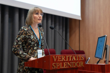 A woman in a patterned dress stands behind a lectern with the inscription “VERITATIS SPLENDOR”. She is wearing a badge around her neck and two microphones are set up in front of her. In the background, curtains and a section of a projection screen are visible. To the right is a monitor with the logo “SAPIENTIAE STUDIUM”.