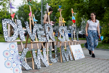 Large letters forming the inscription Diversity x WUM, decorated with mosaic floral patterns. In the background, two people are holding paper flowers. On the right a woman in a white blouse and blue skirt. In the background are trees and fragments of buildings.