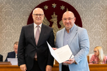 Dwaj mężczyźni stoją obok siebie, trzymając dokument. Jeden ubrany w ciemny garnitur i ma okulary, drugi w jasną marynarkę i koszulę w paski, również ma okulary. W tle ściana z roślinnym wzorem, czerwony herb oraz dwie osoby siedzące przy stole, jedna z laptopem.