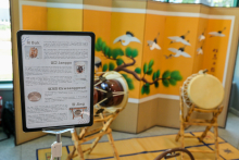 An exhibition of traditional Korean musical instruments in front of an ornate screen with a painting of cranes and pine trees. Visible are a Buk drum, a Janggu drum, a small Kkwaenggwari gong and a large Jing gong. On the left is an information board in Korean and English.