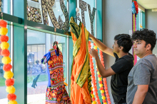 Two men hang colorful outfits on a rack by the window. One holds an orange outfit with green accents. In the background, flower decorations and a mosaic sign. Bicycles and buildings are visible outside the window.