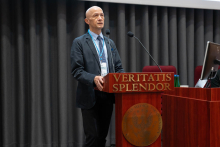 A man, Professor Niemczyk, in a dark suit and blue tie, stands behind a wooden lectern with the inscription “VERITATIS SPLENDOR” and an eagle emblem. On the rostrum are two microphones. Grey curtains in the background, a monitor next to him.