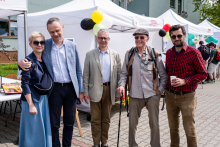 Five people are standing in front of the white exhibition tents. They are the authorities of the Medical Faculty. The two people on the left are elegantly dressed, the third holds a cane and is wearing a light-colored suit. The fourth has a hat and a backpack, the fifth wears a red plaid shirt and holds a mug. In the background are decorative balloons.