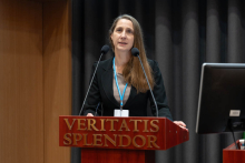 A woman, Prof Domitrz, with dark long hair and in dark formal attire, stands behind a lectern with the inscription “VERITATIS SPLENDOR”. Around her neck is a badge on a blue lanyard. In front of her are two microphones and a computer monitor is visible to her right. The background is made up of dark curtains.