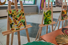 Three easels with paintings in the room. The left painting depicts colorful butterflies on a green background, the middle one - leaves of tropical plants, and the right one - a partially visible floral motif. In the foreground are fans and fabrics, with windows in the background.