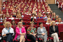 An auditorium filled with attendees seated in red chairs. In the front row, five women in different styles. In the background more rows with people, mostly women. To the right, a staircase leading to the upper rows is visible.