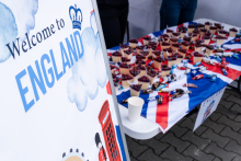 A white banner with the words 'Welcome to England' and a graphic of a cloud, a crown, a red telephone box and a guard. Next to it is a table with the flag of Great Britain, on which lie paper cones with sweets and souvenirs.