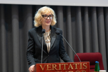 A woman, the Deputy Minister of Health, with fair hair, wearing glasses and a dark suit and striped shirt, stands behind a lectern with the inscription “VERITATIS SPLENDOR”. She speaks, smiling gently. In front of her are two microphones, next to her a bottle of water and a glass. In the background, grey curtains and a fragment of a red chair.