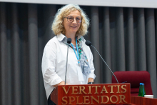 A woman, the pro-dean, with light, curly hair and wearing glasses, stands behind a lectern with the inscription “VERITATIS SPLENDOR”. She is dressed in a white shirt and coloured scarf. In front of her are two microphones. Dark curtains are visible in the background. To the right, on a table next to the lectern, stands a water bottle and a cup.