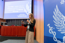 A woman stands on stage and holds a microphone. In the background is a screen with a presentation entitled “Stroke - a clinical and social problem in the context of current recommendations - the role of a clinical speech therapist”. Under the title are the details of the speaker: Izabela Domitrz, Department of Neurology, WUM, Bielański Hospital in Warsaw. To the right of the screen is the logo with Aesculapius' cane.