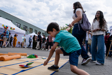 A boy in a green T-shirt and blue shorts throws colored bags on a mat laid out on the pavement. In the background, a group of people and stands with white tents, on one of which colorful balloons are visible.
