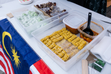 A table with dishes and the Malaysian flag. On the table three containers: green balls with white powder, pieces of chocolate cake and yellow pancakes with a bowl of red sauce and a ladle. Nearby lie wooden chopsticks and plastic cutlery.