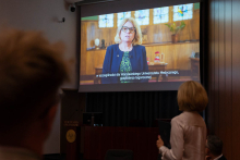  Sala konferencyjna z dużym ekranem, na którym wyświetlany jest film z kobietą prof. Bogumiła Kaniewska, przewodnicząca KRASP. Kobieta ma na sobie granatową marynarkę i zieloną bluzkę. Przemawia.