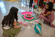 Two women in traditional Indian dress sit on the floor and create rangoli - a decoration made of colored powder in the shape of a flower with pink petals, a yellow center and white patterns. Containers of powder and tools lie around.