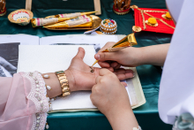 Two people participate in painting their hands with henna. The person on the left in traditional costume with a bracelet holds the hand on which the other person applies henna with a gold tube. On the table lie henna cones, colorful boxes and ornaments.