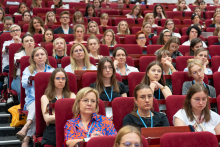 Auditorium with a large group of people seated in red chairs, facing the stage out of frame. Participants have badges on lanyards.