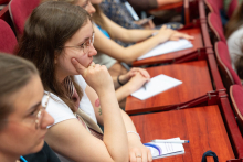 A group of people sit in rows in the lecture theatre. Some of them are taking notes on paper. Wooden table tops and red chairs are visible.