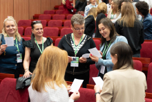„Grupa osób stoi w sali konferencyjnej z czerwonymi fotelami, prowadząc rozmowy i trzymając kartki. Większość ma identyfikatory na zielonych smyczach. W tle inne osoby rozmawiają lub przemieszczają się.