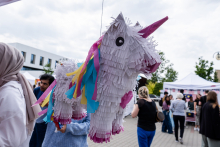 Pinata in the shape of a unicorn suspended on a string. Made of white paper, with pink horn and hooves and mane and tail in blue, yellow and pink. In the background are participants of the outdoor event, tents and buildings.