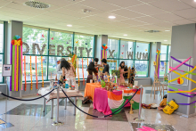 Colorfully decorated indoor space with two tables full of decorations, including the flag of India. In the background, people preparing the display. On the windows the sign “Diveristy x WUM” decorated with flowers and garlands. The room is surrounded by railings, tile floor.