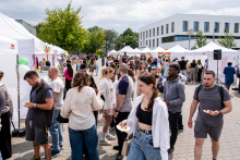 A crowd of people at an outdoor event. In the background are rows of white exhibition tents and a modern building with large windows. Attendees dressed variously, some holding plates of food. The event takes place on a paved square surrounded by greenery. The weather is cloudy, but bright.